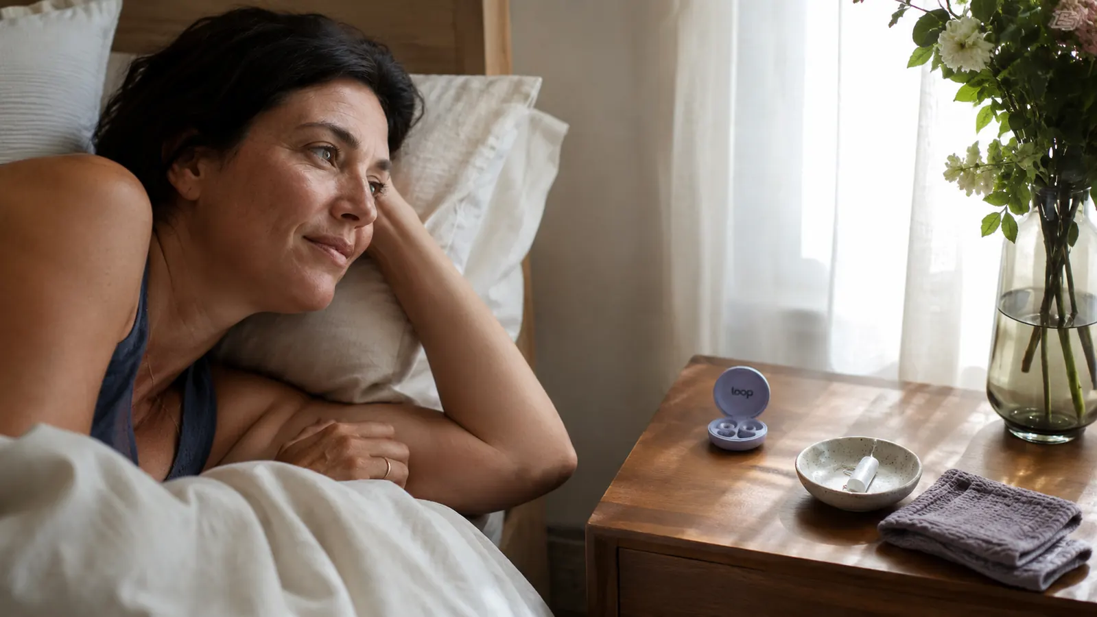 Bedroom nightstand scene with earplug case, small dish, and folded washcloth, morning light through curtains